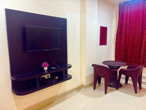 Una habitación con un televisor de pantalla plana montado en una pared de madera oscura, una pequeña mesa con dos sillas y una cortina roja.