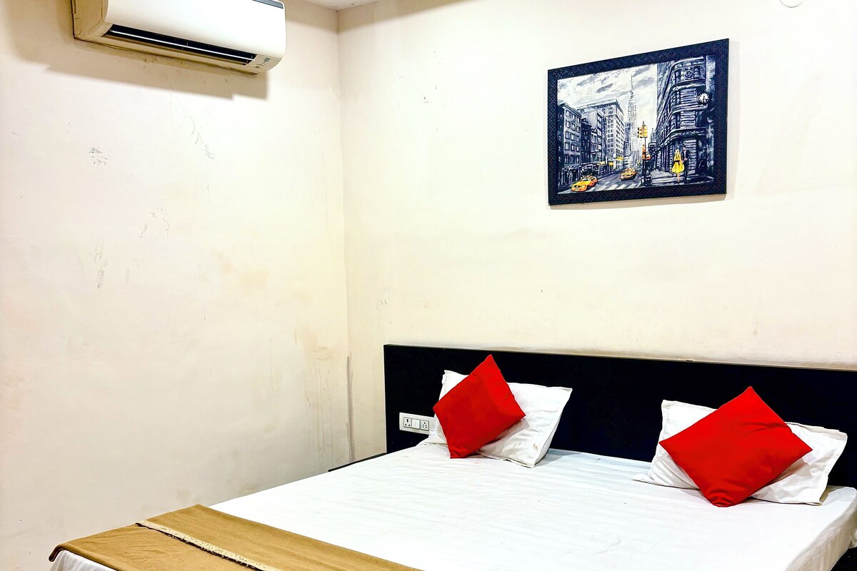 Habitación de hotel con cama, almohada roja y blanca, manta beige, alfombra estampada, unidad de aire acondicionado y una pintura de un paisaje urbano en la pared.