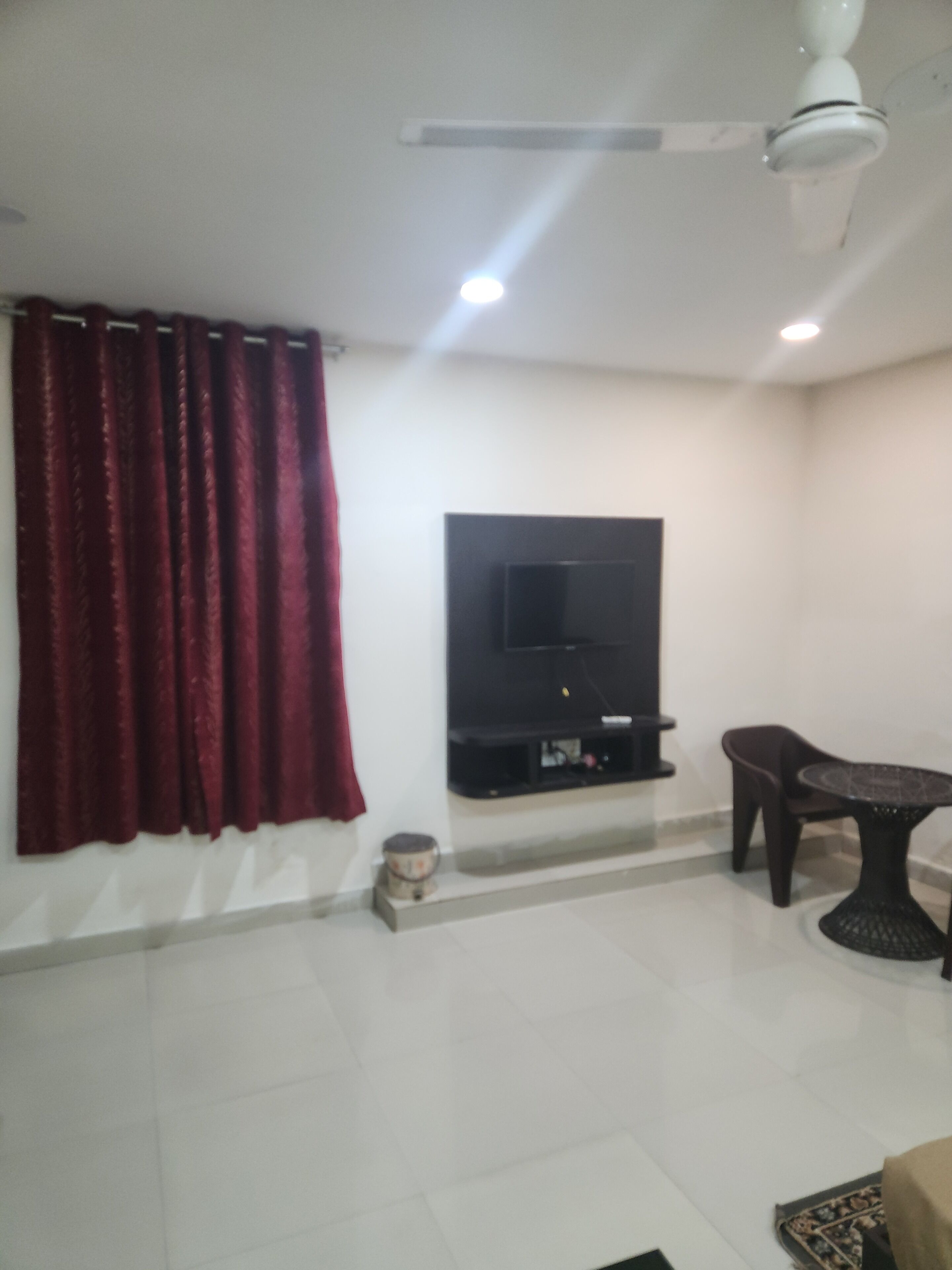 A living room with a flat-screen TV mounted on the wall, a ceiling fan, a dining table with chairs, and a red curtain.