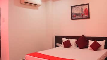 Un dormitorio con cama, almohadas, mesita de noche, una silla, un equipo de aire acondicionado, ventilador de techo y un cuadro en la pared.