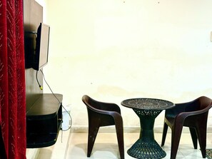Una habitación con un televisor montado en la pared, una mesita redonda con base tejida y dos sillas de plástico.