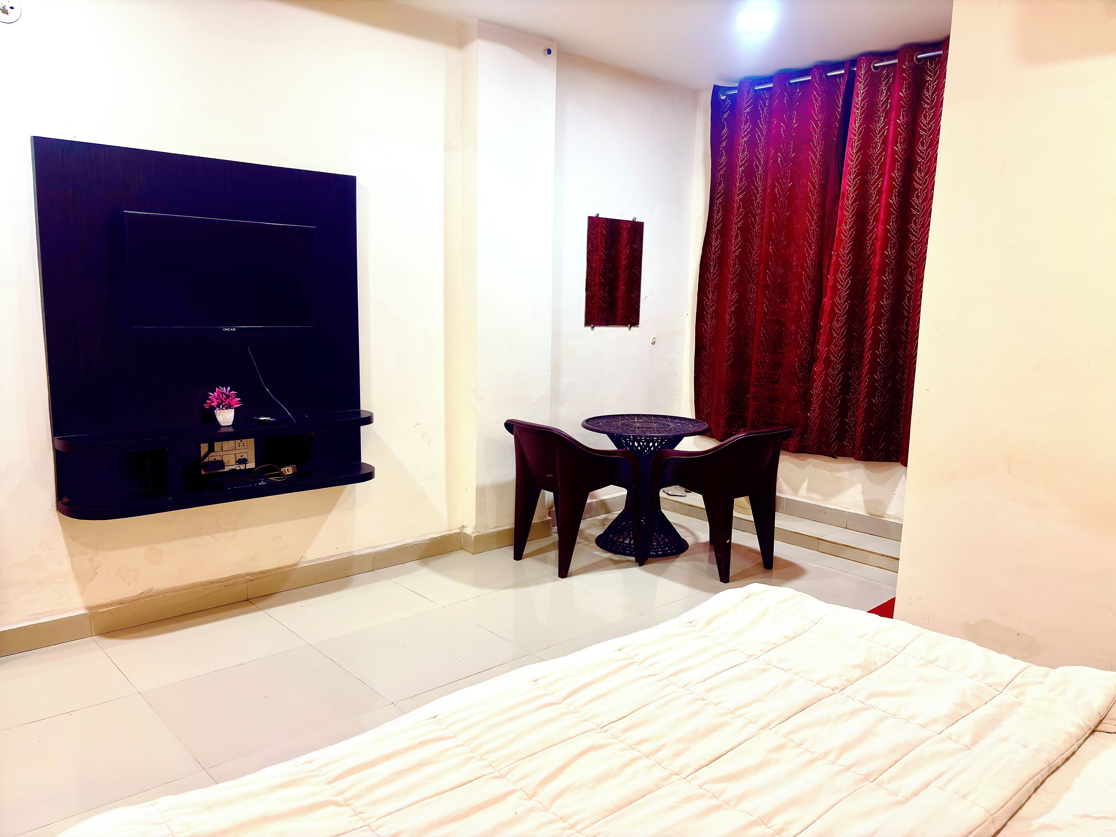 A room with a bed, a small table with two chairs, a TV mounted on the wall, and a window with red curtains.