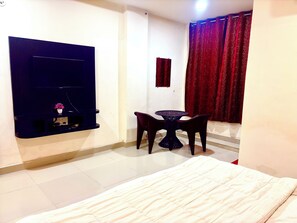 A room with a bed, a small table with two chairs, a TV mounted on the wall, and a window with red curtains. - HOTEL SAI KRIPA  (Huzur)
