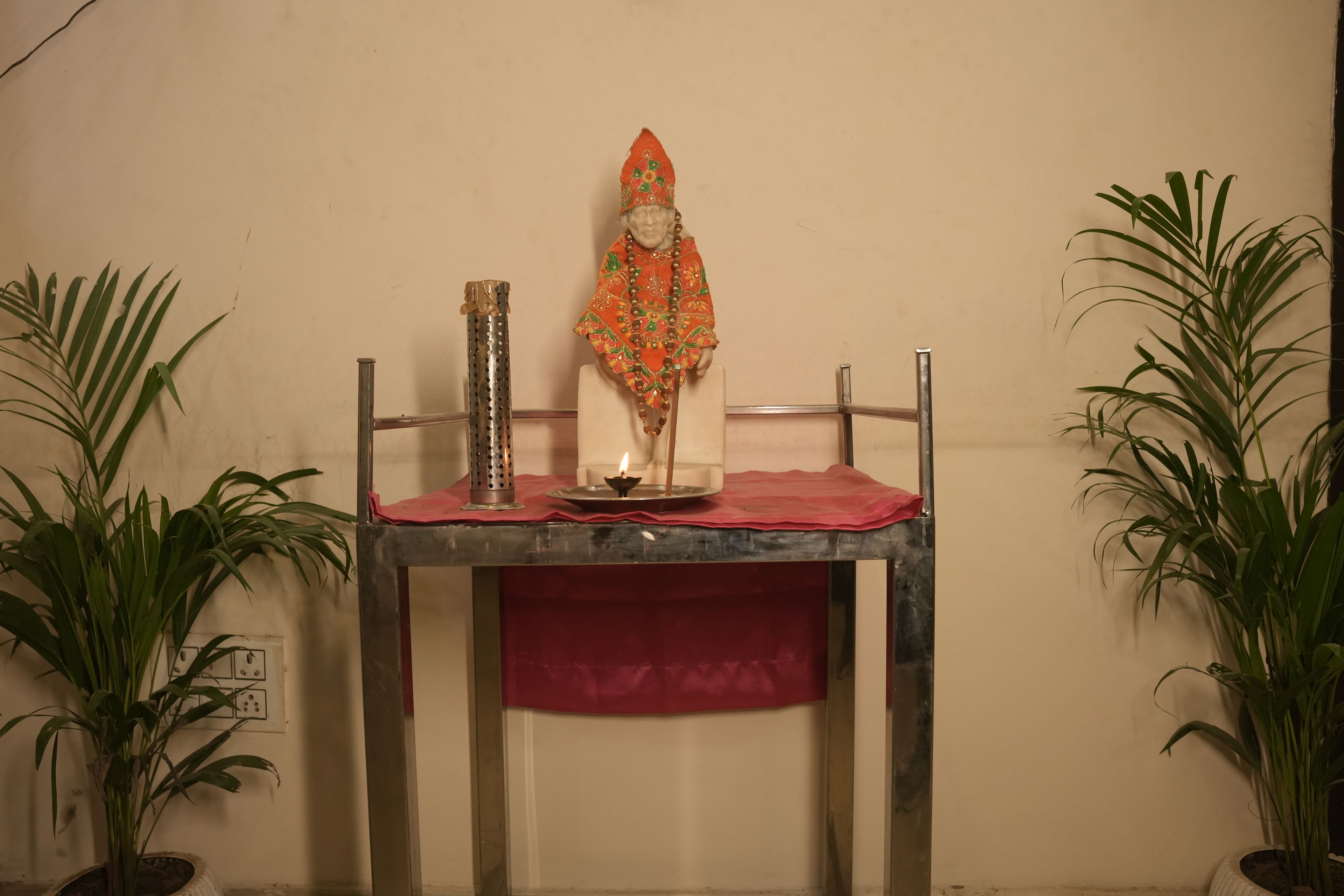 Una mesa con una estatua, un recipiente metálico y una vela encendida. Hay dos plantas en macetas a cada lado de la mesa.