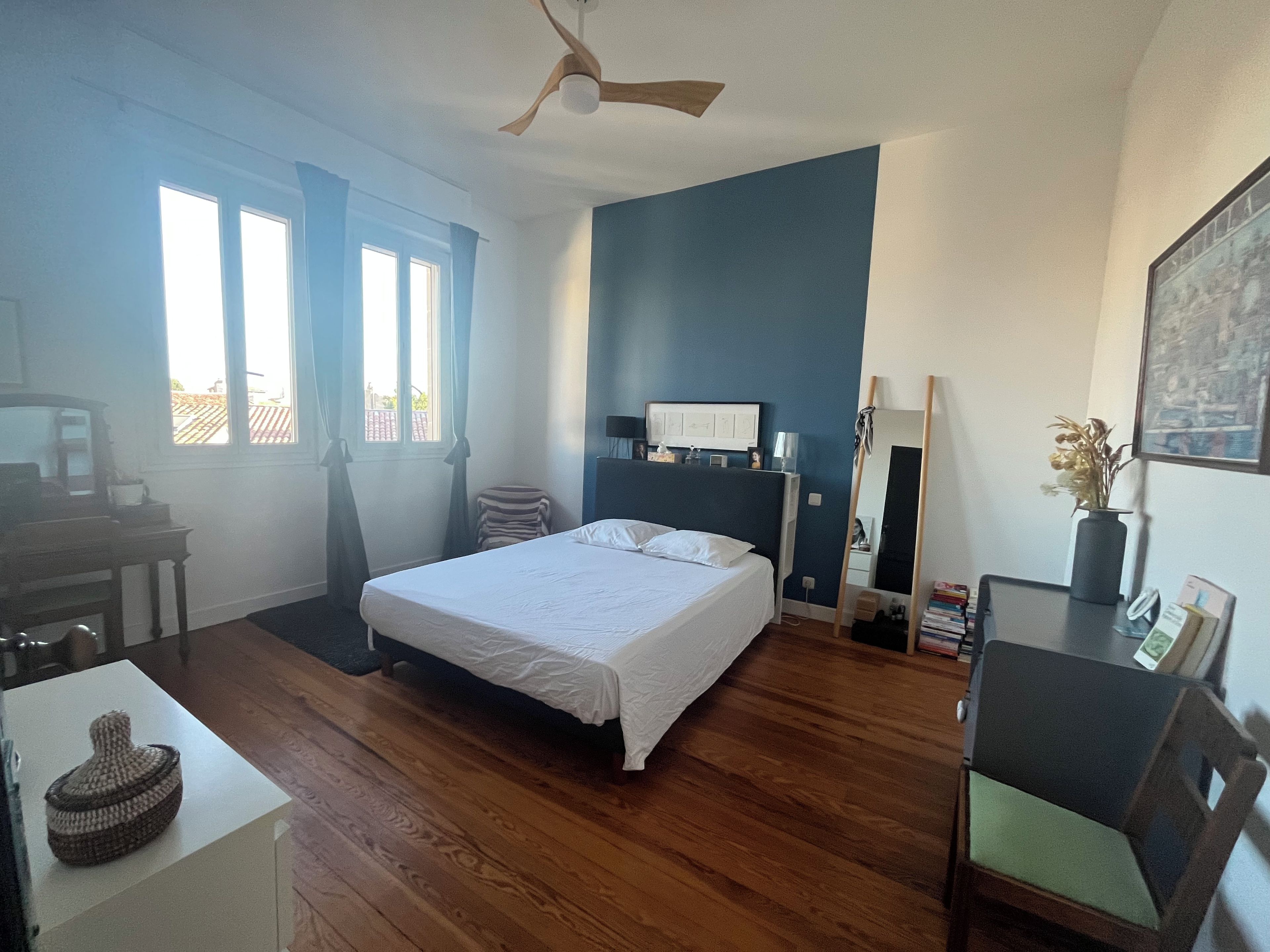 A bedroom with a bed, a desk, a chair, a ceiling fan, a window with curtains, and a wall-mounted mirror.