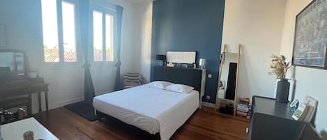 A bedroom with a bed, a desk, a chair, a ceiling fan, a window with curtains, and a wall-mounted mirror.