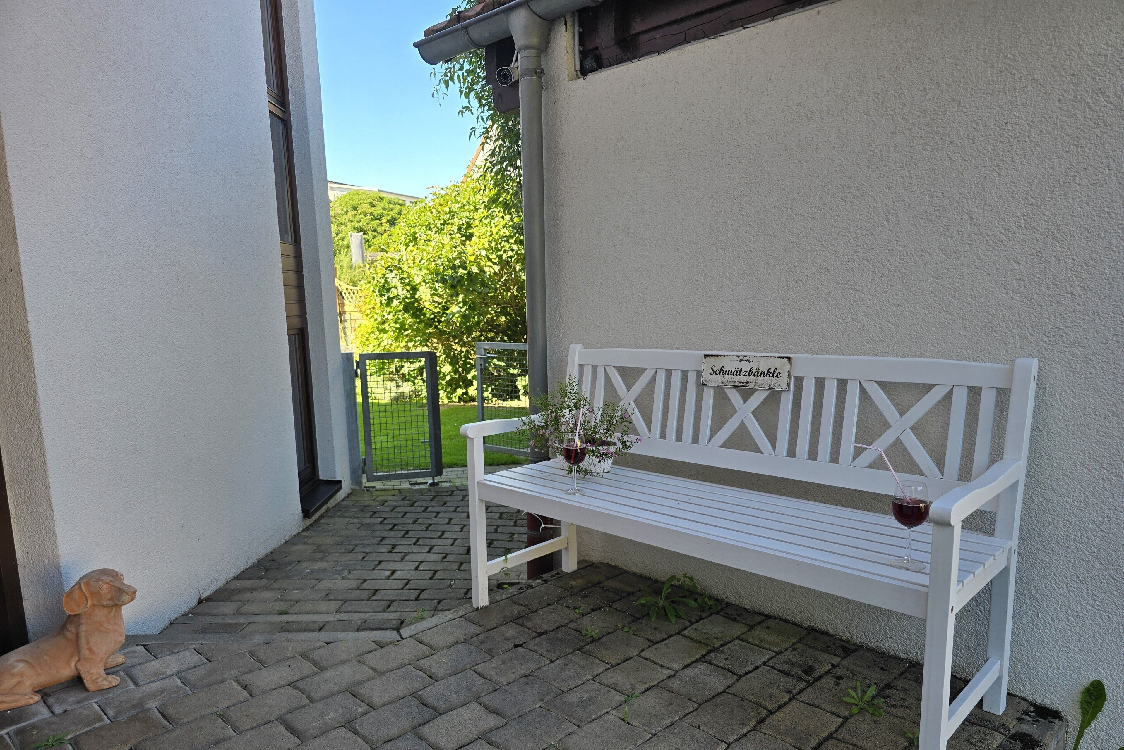 Eine weiße Bank mit einem Schild, einer kleinen Hund-Statue und einem Blumentopf auf einer gepflasterten Terrasse.