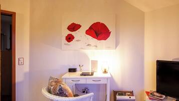 Une pièce avec un bureau blanc, une chaise en osier, un téléviseur à écran plat et une œuvre d’art représentant des fleurs rouges, accrochée au mur.