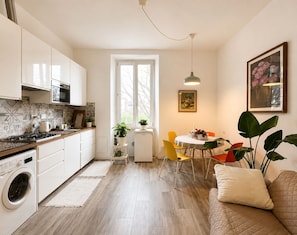 A modern kitchen with a stove, oven, and washing machine, a dining area with a table and chairs, and a living area with a sofa and a plant. - Room-natures Retreat - Private Room - N (Florence)