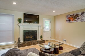 A living room with a fireplace, a flat-screen TV mounted above it, a modern coffee table, and a contemporary abstract painting on the wall. - Kennesaw Home Near KSU & Lake Acworth (Kennesaw)
