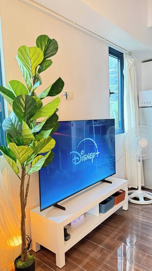 A flat-screen TV on a white stand with a Disney+ logo, a potted plant, and a standing fan in a room with a window and curtains.