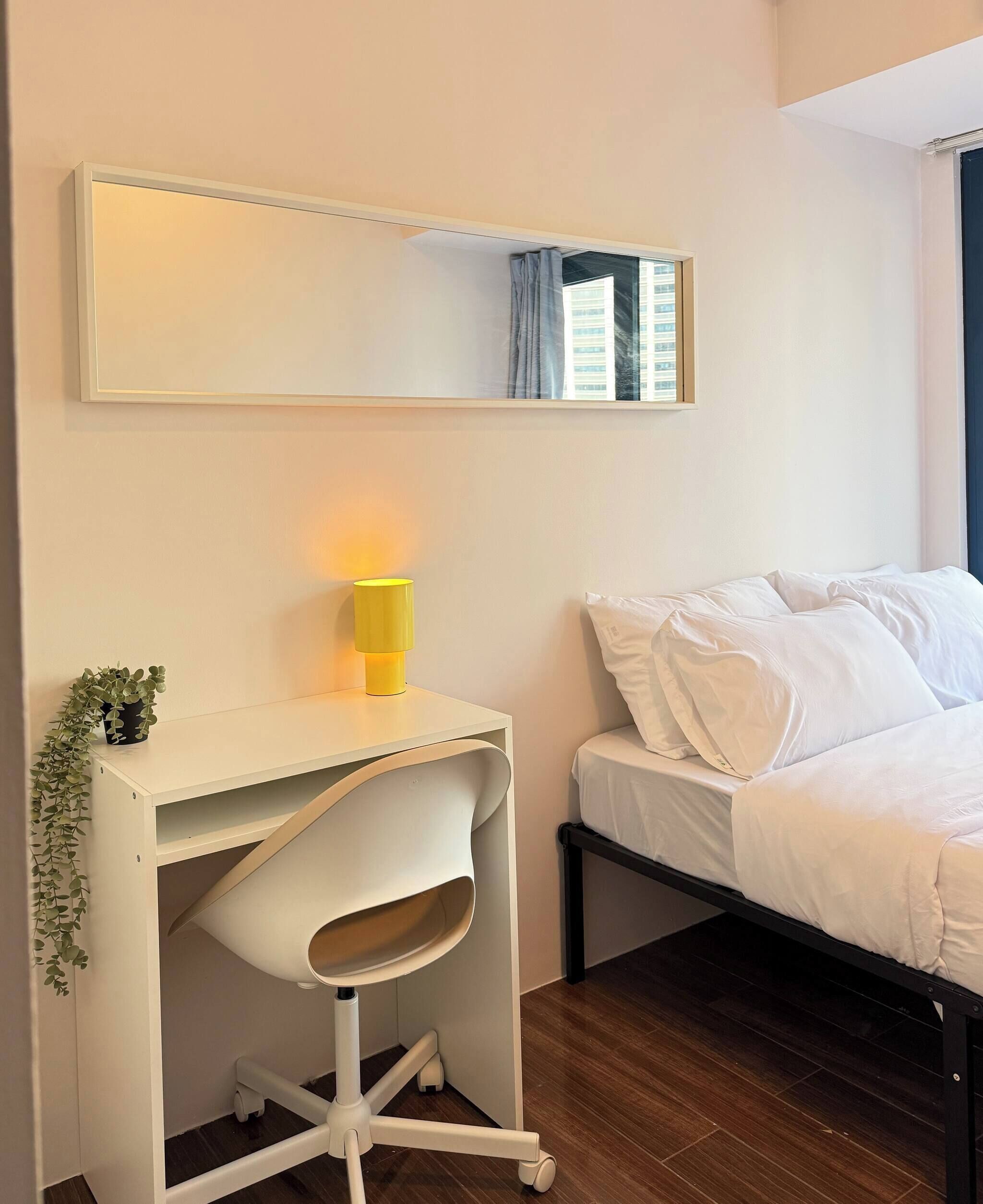 A bedroom with a bed, a desk with a chair, a small plant, and a mirror on the wall.