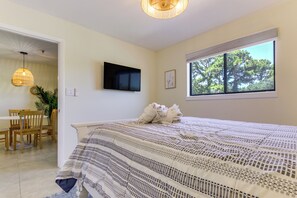 A bedroom with a bed, a television mounted on the wall, a window with a view of trees, and a dining area with chairs and a table. - Relax, Recharge, Repeat: Hilton Head Resort Condo (Hilton Head Island)