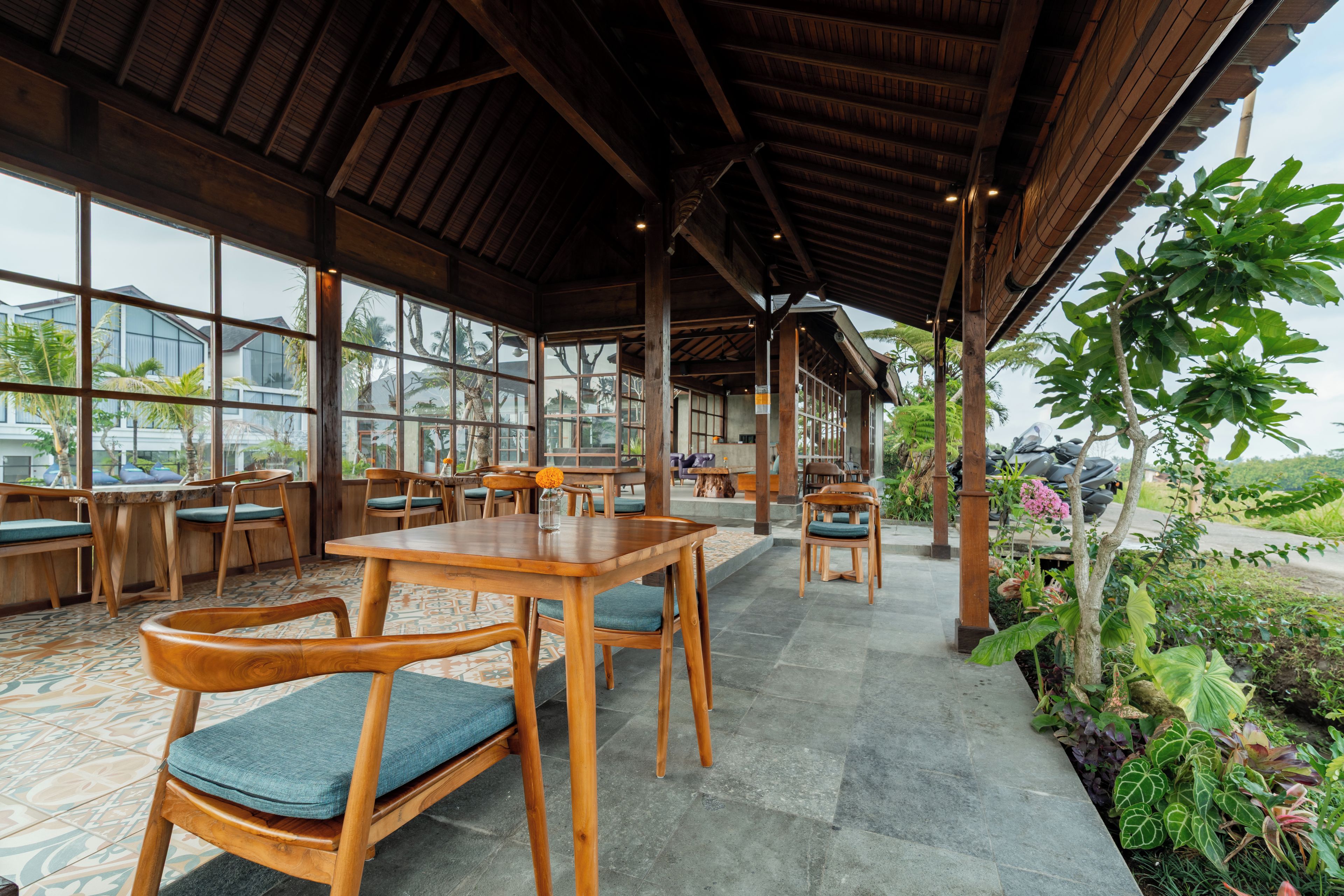 A covered outdoor seating area with wooden tables and chairs, large windows, and a view of the surrounding landscape.