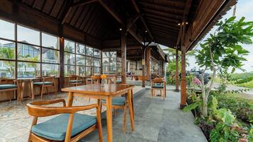 A covered outdoor seating area with wooden tables and chairs, large windows, and a view of the surrounding landscape.