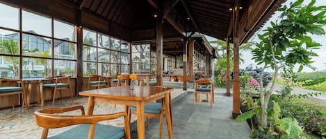 A covered outdoor seating area with wooden tables and chairs, large windows, and a view of the surrounding landscape.