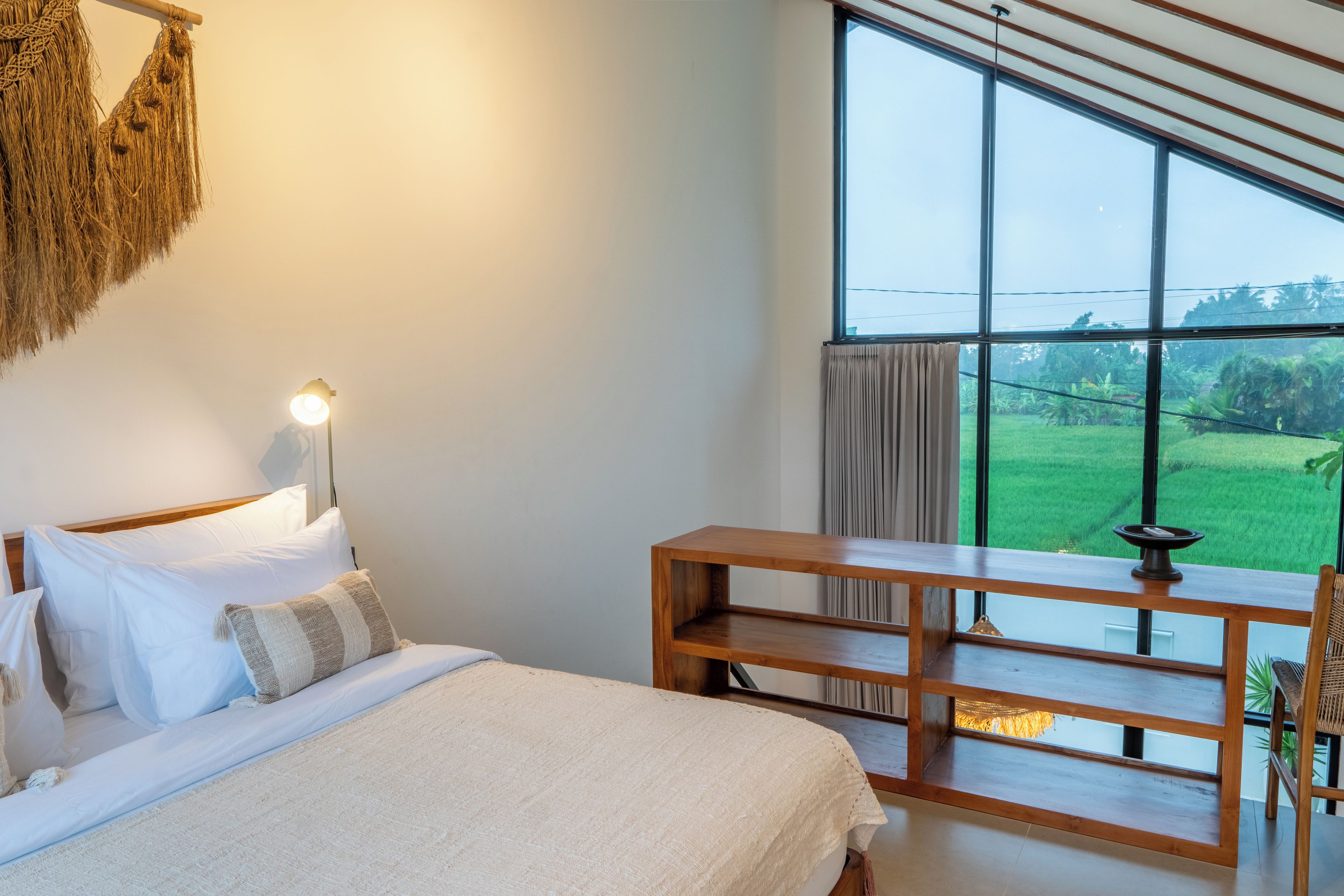 Ein Schlafzimmer mit großem Fenster, einem Bett mit weißer Bettwäsche, einer Nachttischlampe und einem Holfnachtkästchen.