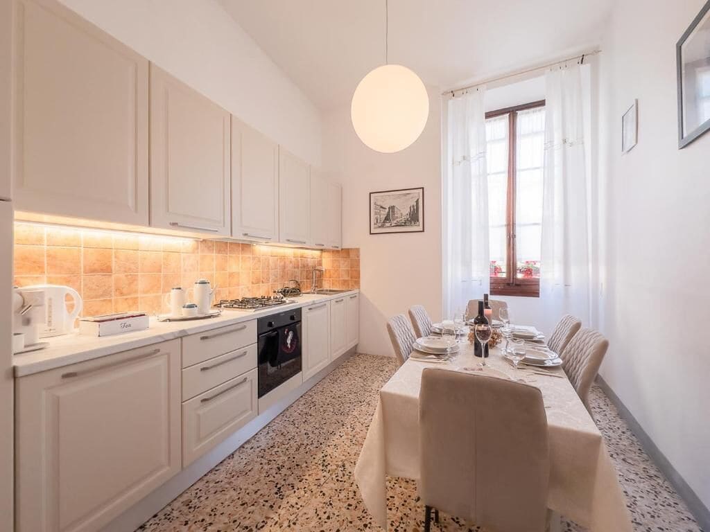 Une cuisine moderne avec des meubles blancs, une table à manger dressée pour six personnes et une grande fenêtre agrémentée de rideaux.