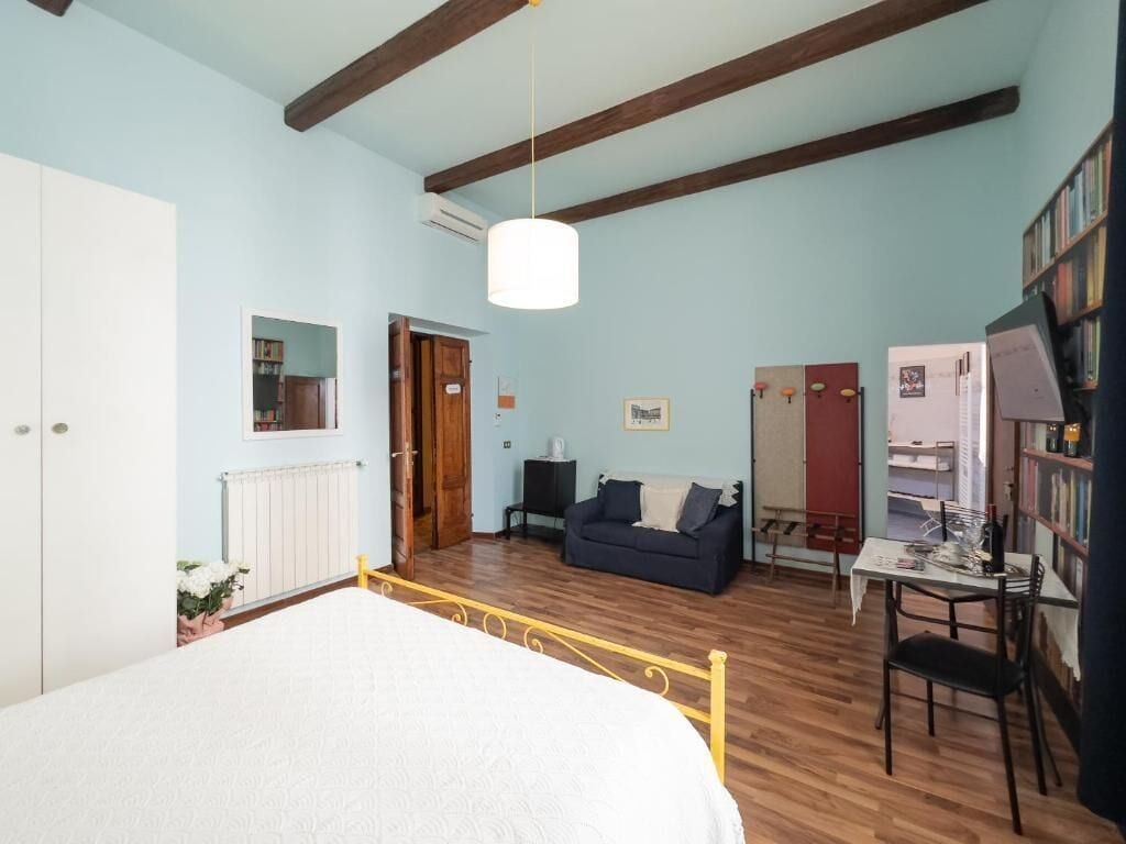 A bedroom with a bed, a wooden floor, a white radiator, a blue wall, a wooden ceiling, a white door, a wooden wardrobe, a white bedside table, a white lamp, a blue sofa, a wooden table, a chair, and a bookshelf.