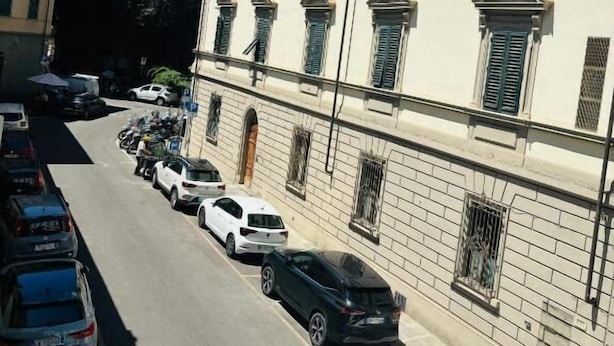 A street view with parked cars and a building with multiple windows and shutters.