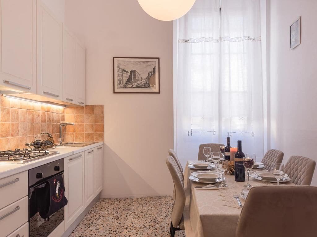 A compact kitchen with a gas stove, oven, and sink, a dining table set for two with chairs, and a window with sheer curtains.
