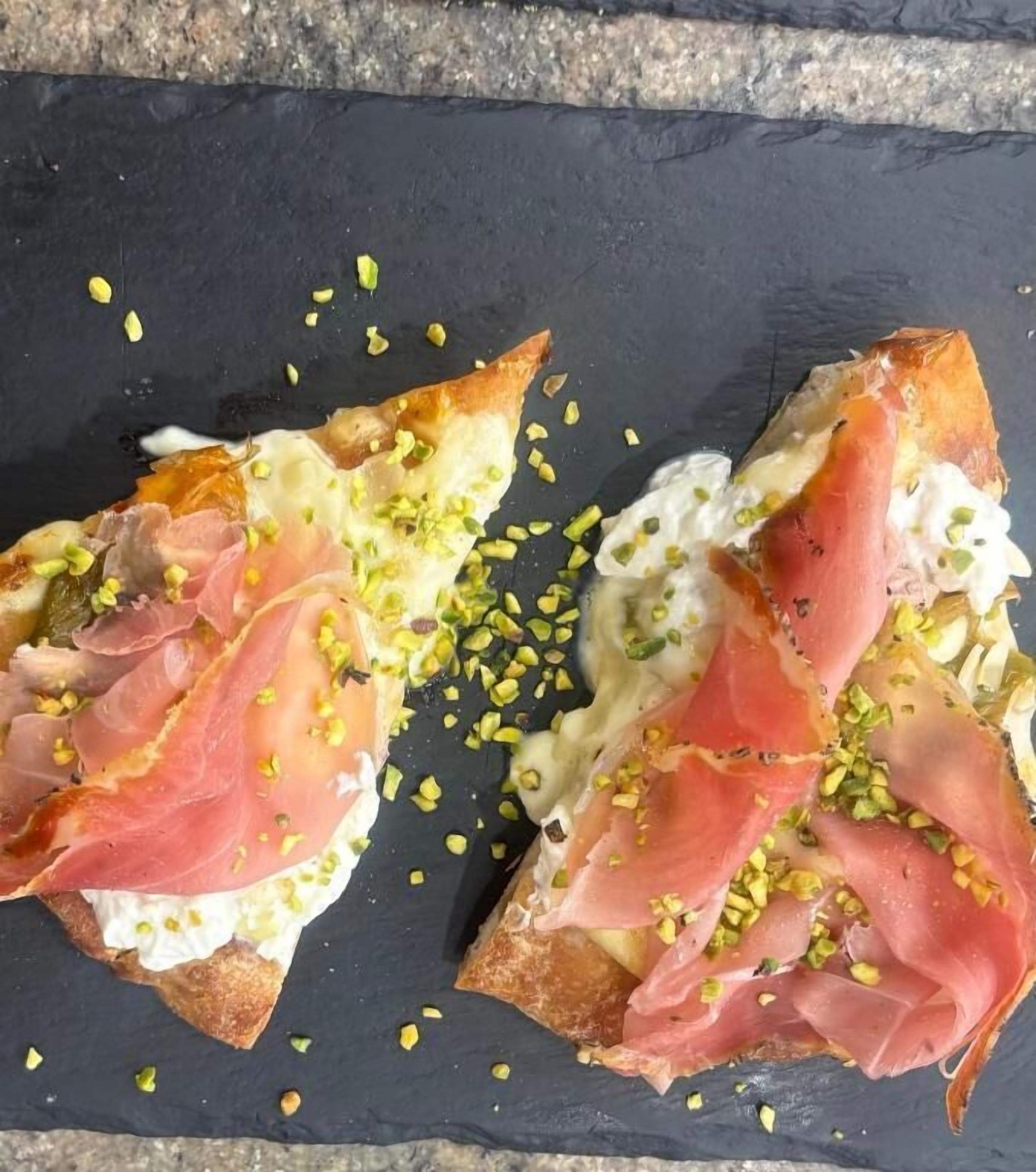 A black slate serving board with two pieces of bruschetta topped with prosciutto and a creamy white spread, garnished with chopped green herbs.