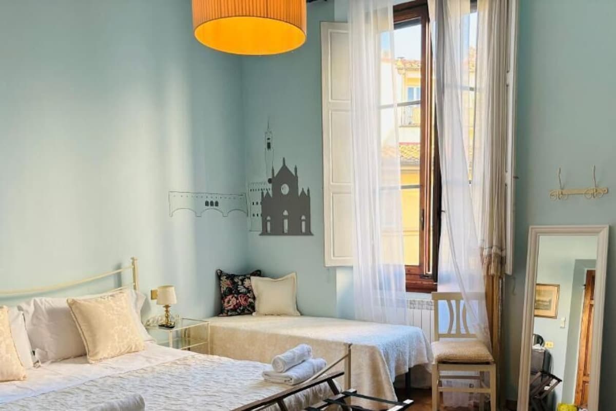 A bedroom with two beds, a window with curtains, a mirror, and a wall-mounted artwork.