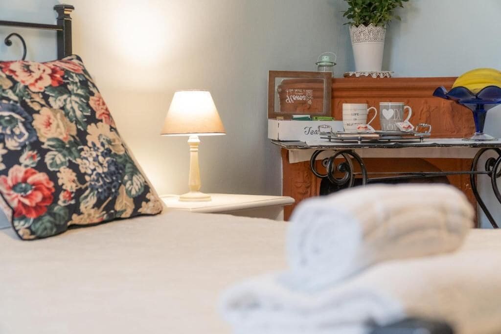 Ein Bett mit einem blumigen Kopfkissen, ein Nachttisch mit Lampe, eine kleine Holzablage mit Pflanze und ein gerahmtes Bild.