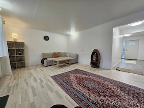 A living room with a sofa, coffee table, and a large rug. There is a clock on the wall and a lamp on a shelf.