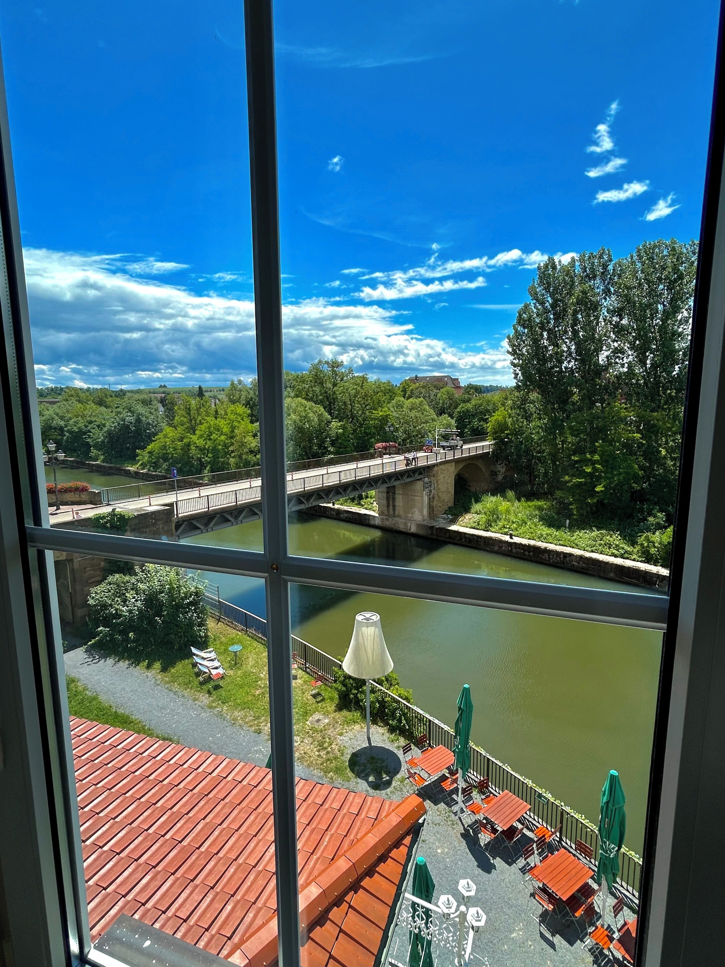 Blick auf einen Fluss mit einer Brücke, einer Außenbestuhlung und Grünflächen.