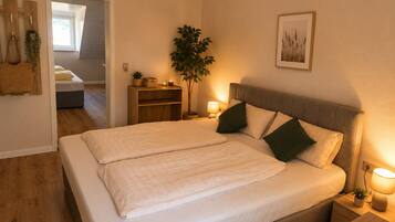 Een tweepersoonsbed met hoofdeinde, nachtkastjes met lampen, een plantje op een nachtkastje, een houten plank met een plant, en een lijst met een afbeelding aan de muur.