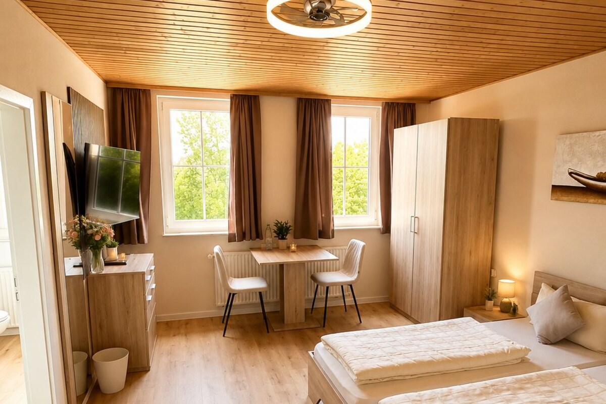 A room with a wooden ceiling, a bed, a desk with chairs, a television, and a window with curtains.
