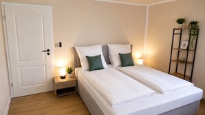 A bedroom with a double bed, bedside tables, a nightstand, a door, and a wall-mounted shelf with plants.