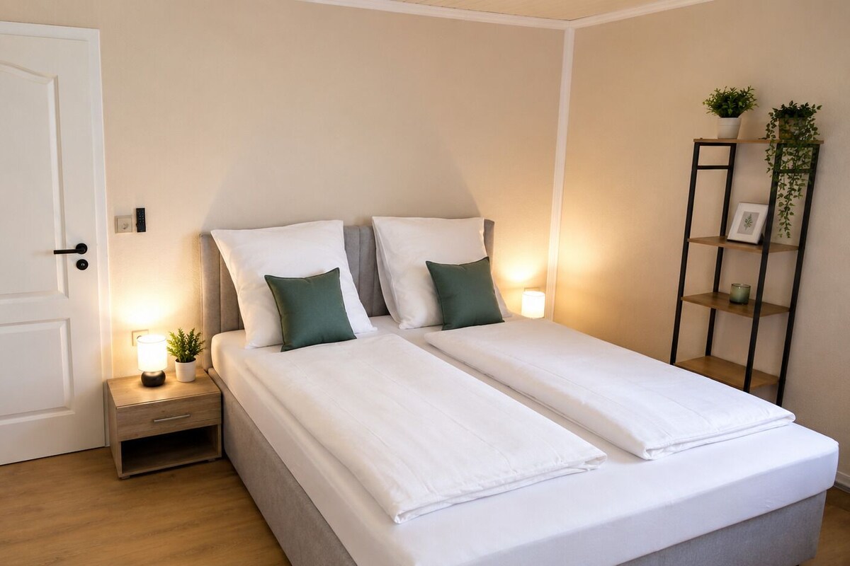 A bedroom with a double bed, bedside tables, a nightstand, a door, and a wall-mounted shelf with plants.