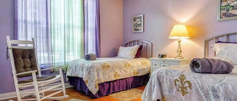 Un dormitorio con dos camas, una mecedora, una mesita de noche con lámpara y una ventana con cortinas.