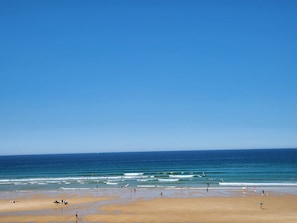 Una playa con gente y tablas de surf, cielos despejados y olas marinas tranquilas.