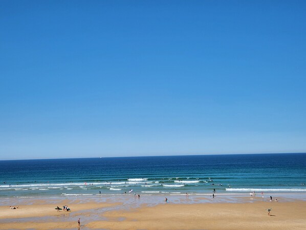 Una playa con gente y tablas de surf, cielos despejados y olas marinas tranquilas.