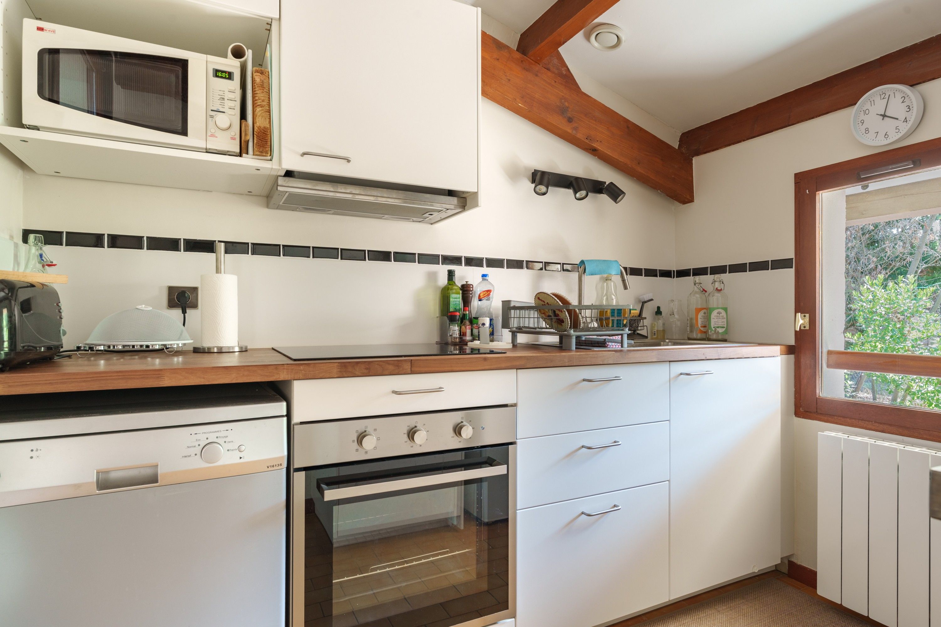 A kitchen with white cabinets, a microwave, a stove, and a sink. There is a clock on the wall and a window with a view of greenery.