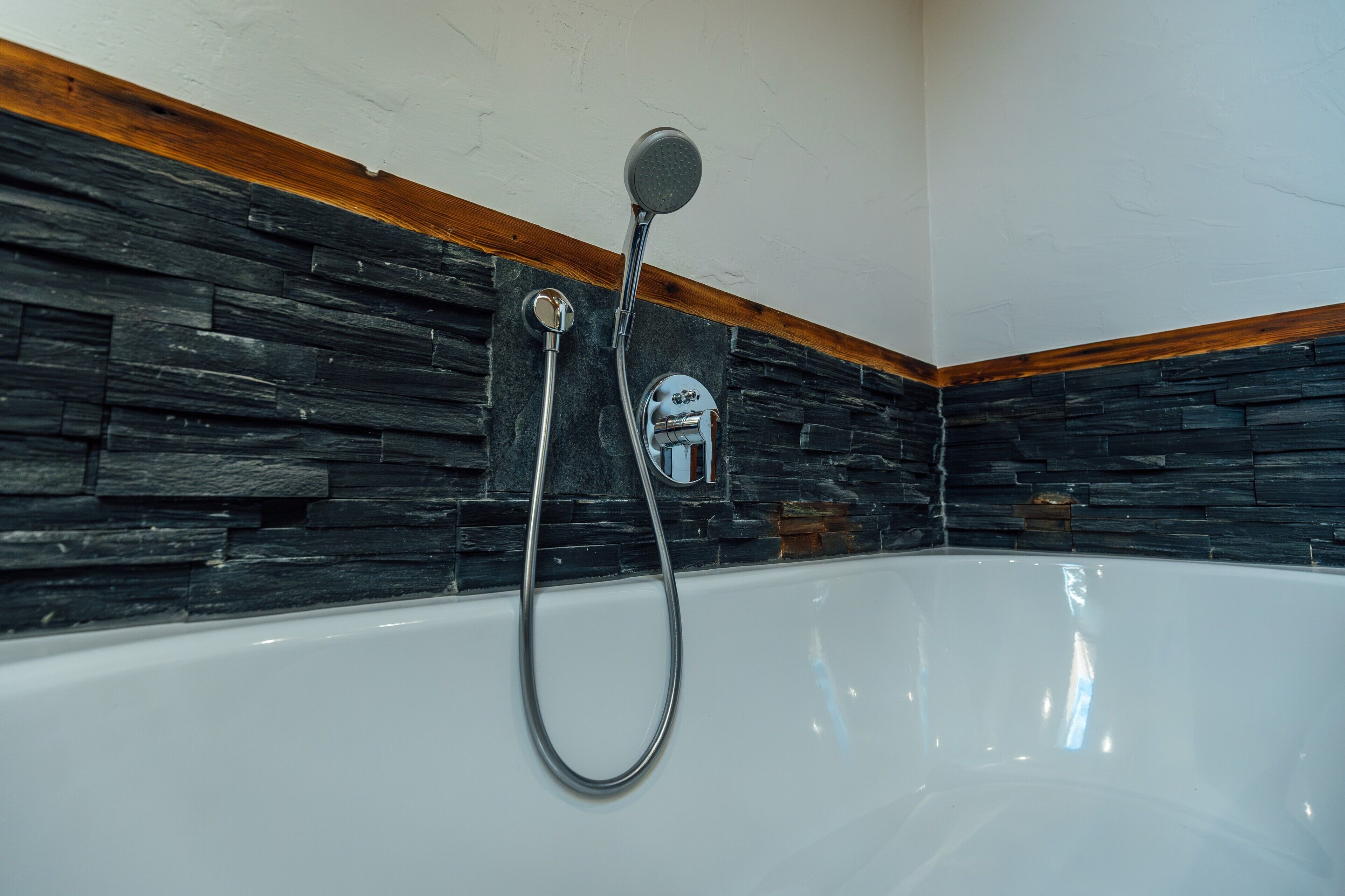 Eine Badewanne mit Duschkopf und Brause, vor einer Steinwand mit Holzverkleidung.