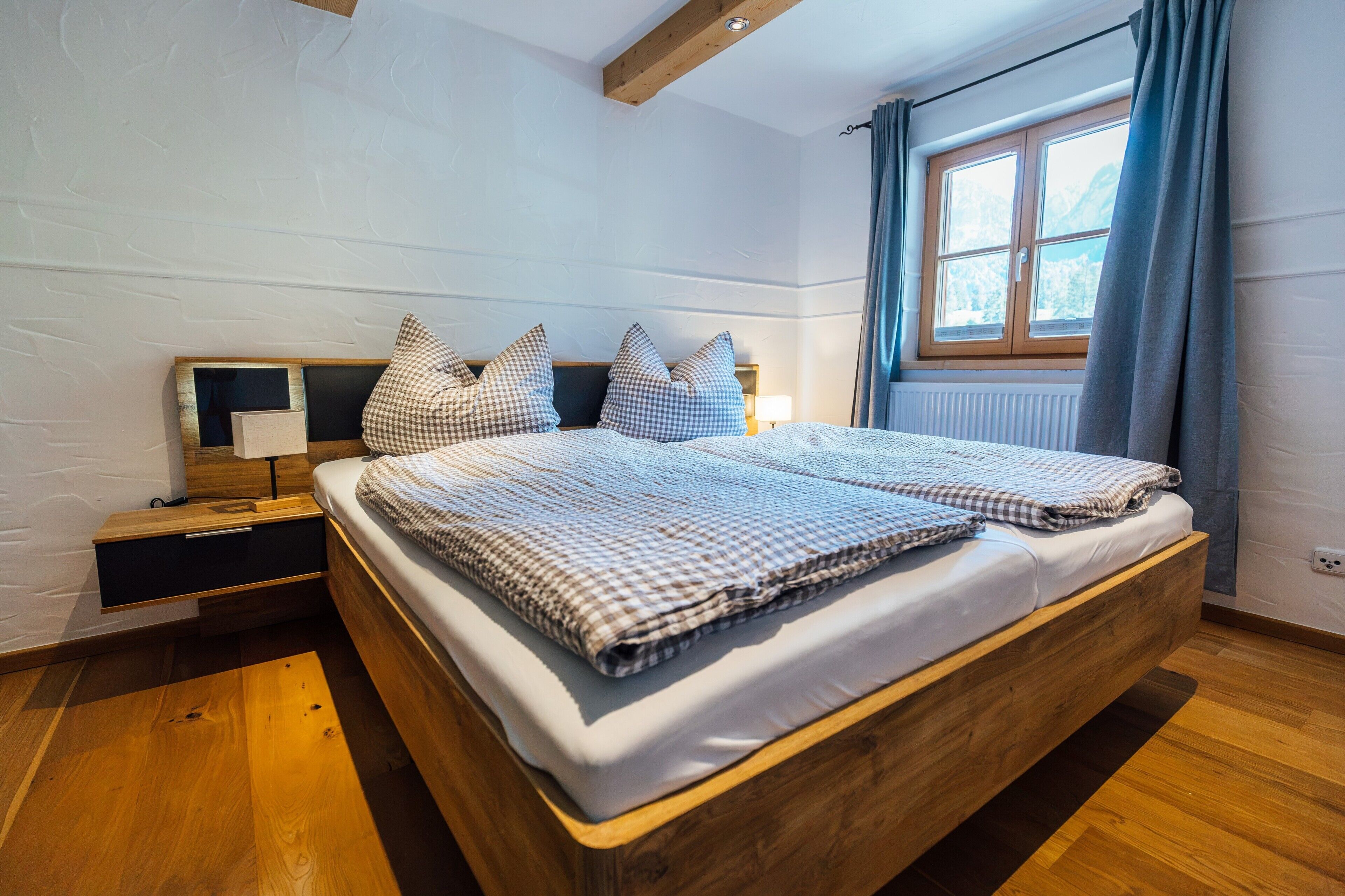 Ein Schlafzimmer mit einem hölzernen Bett, karierter Bettwäsche, einem Nachttisch, einer Lampe und einem Fenster mit Vorhängen.
