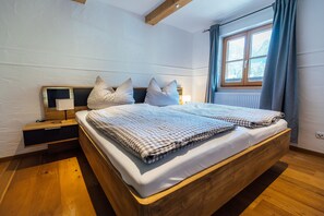 Un dormitori amb llit de fusta, roba de llit a quadres, una tauleta de nit, una làmpada i una finestra amb cortines.