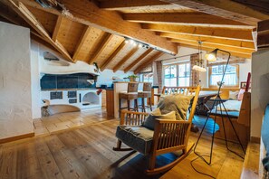 A cozy room with wooden beams, a stone fireplace, a rocking chair, and a dining area with a table and chairs.