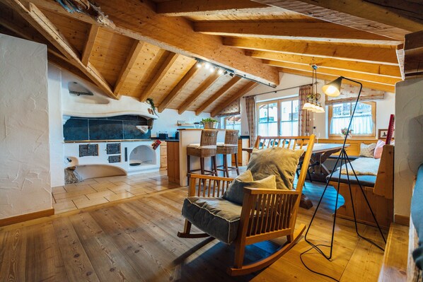 Una habitación acogedora con vigas de madera, chimenea de piedra, una mecedora y un comedor con mesa y sillas.