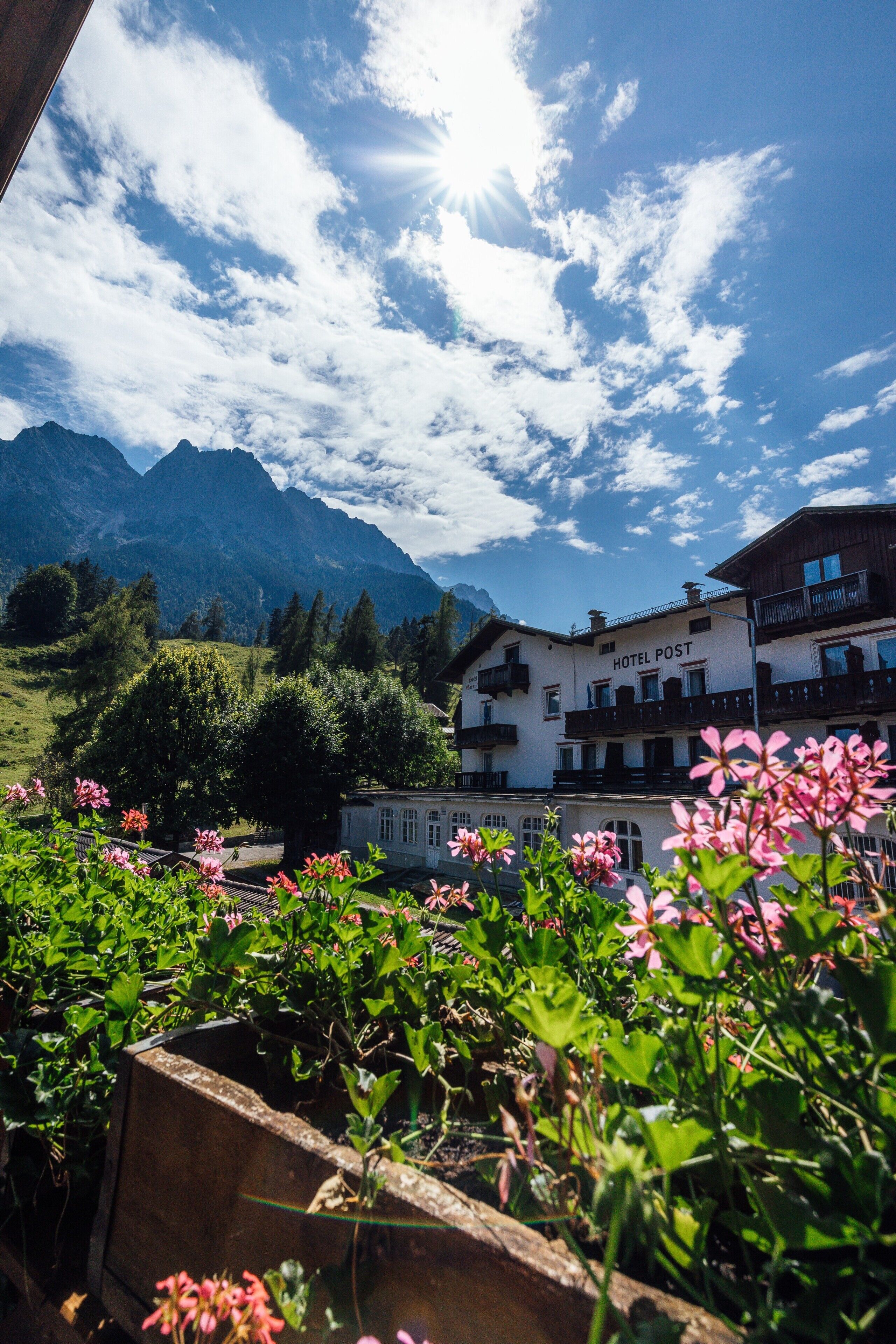 A hotel with a mountain backdrop, a garden with pink flowers, and a clear sky.