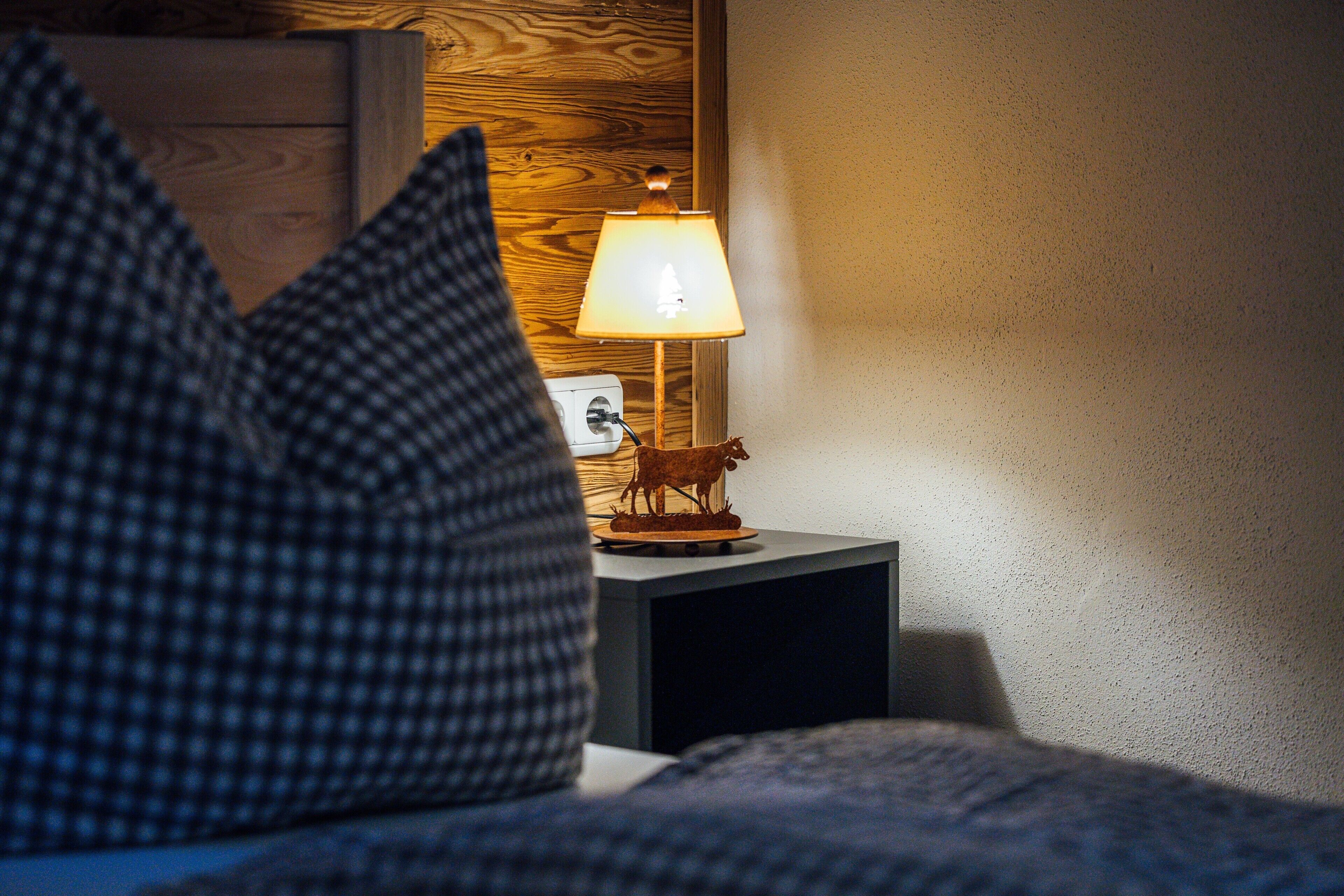 Ein Bett mit einem karierten Kopfkissen, einer Nachttischlampe mit einer Kuhfigur und einem hölzernen Kopfteil.