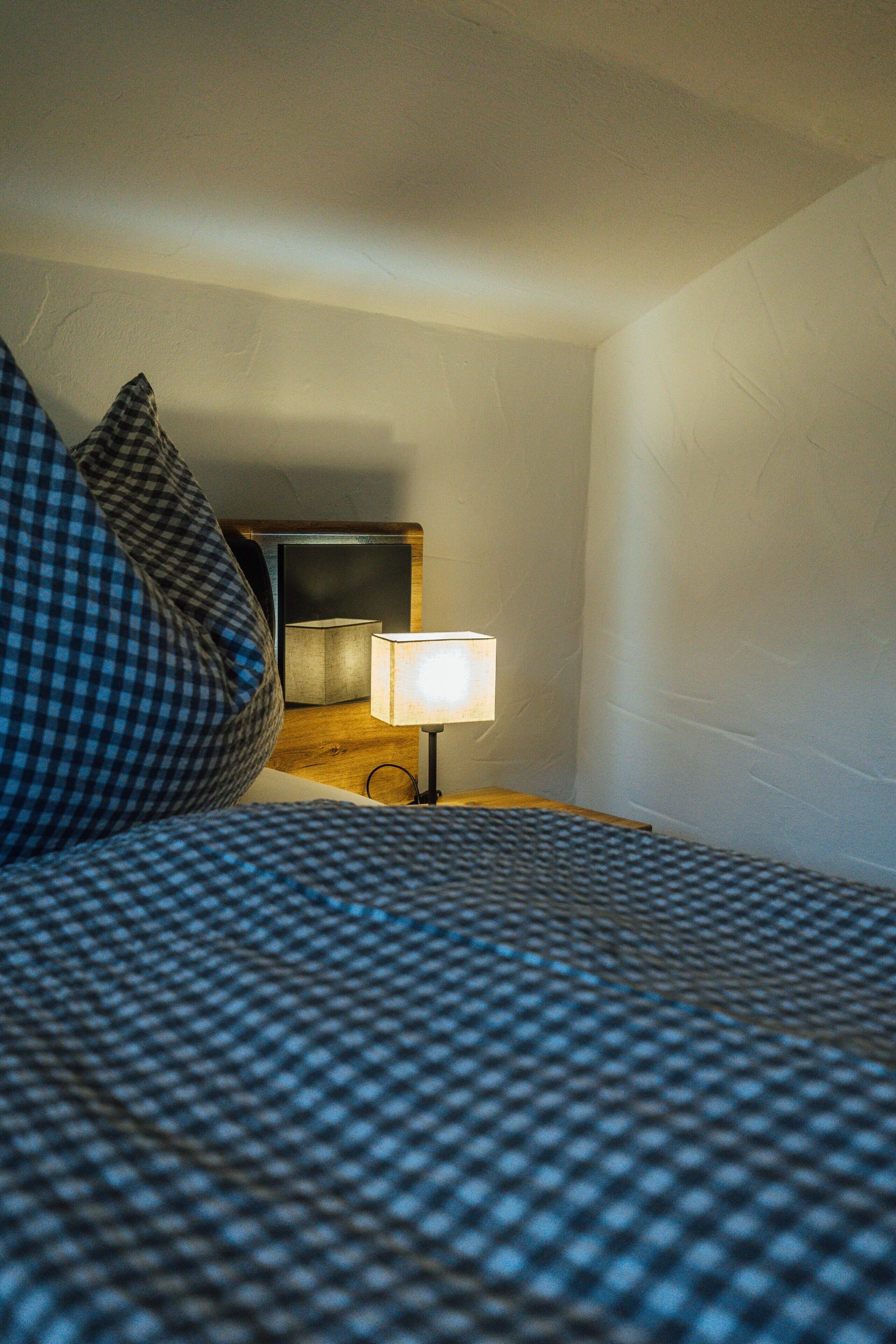 Een bed met een geruit sprei en een kussen, een nachtkastje met een lamp, en een spiegel aan de muur.