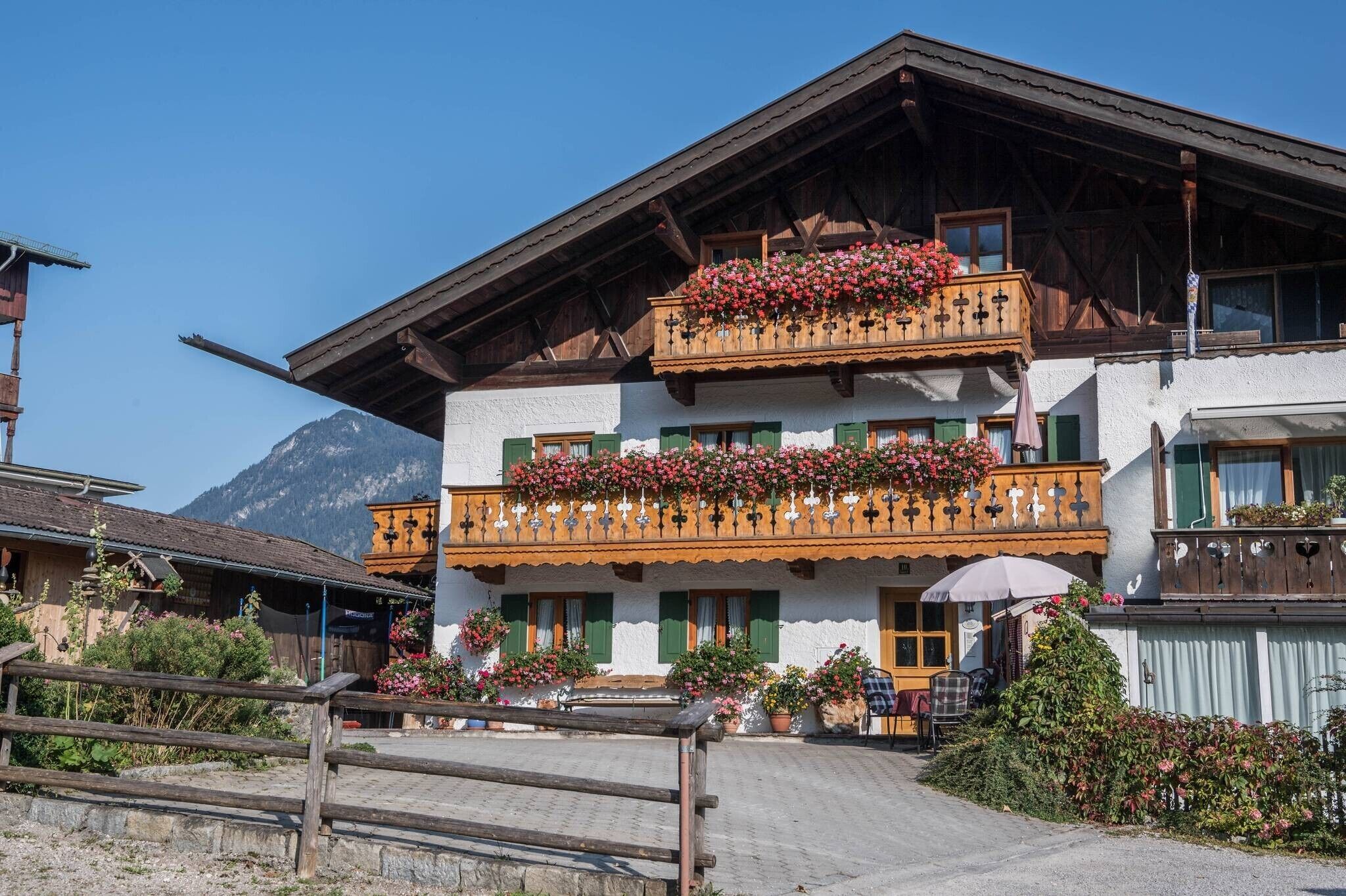 A traditional chalet with a wooden balcony adorned with flowers, a mountain backdrop, and a paved entrance.
