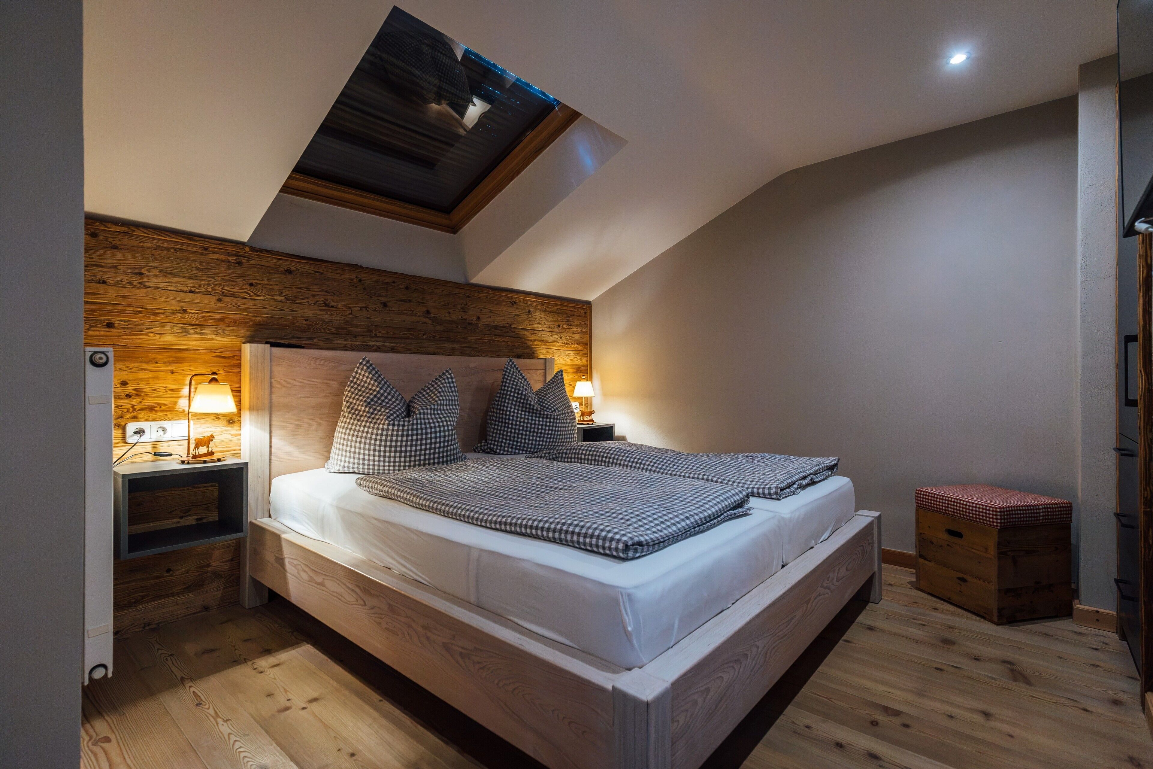 Un dormitorio con techo inclinado, cabecera de madera, cama con ropa de cuadros, lámparas de noche y un pequeño cofre de madera.