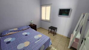 A bedroom with a bed, a nightstand, a small table, a window with blinds, and a picture on the wall.
