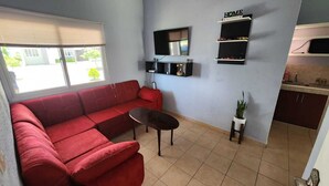 Uma sala de estar com um sofá vermelho, uma mesinha e uma TV fixada na parede.